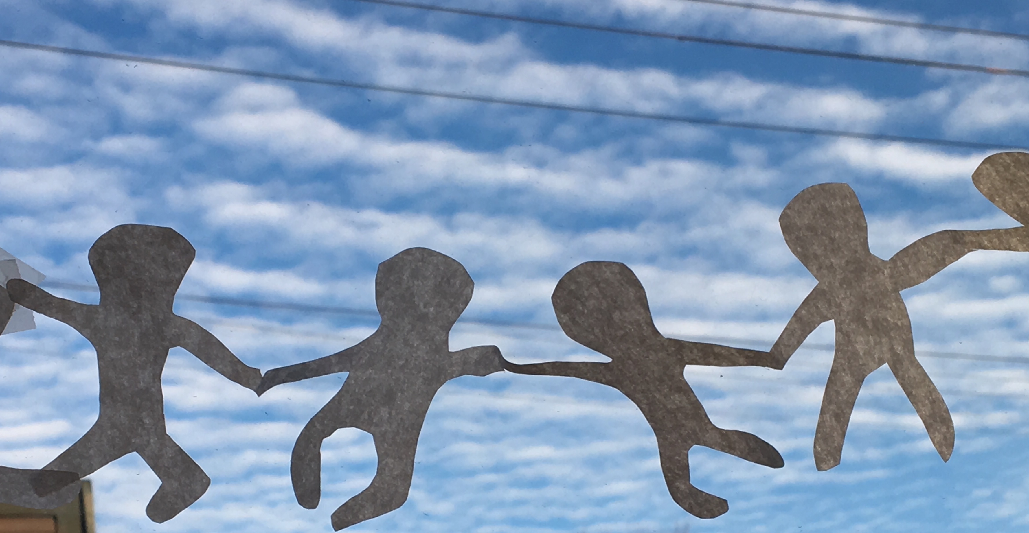 Chain of paper dolls cut from a paper napkin, hanging in a window, with blue sky and powerlines behind